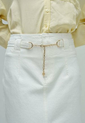 Primer plano de pantalones blancos de tiro alto con trabillas unidas por una delicada cadena dorada y una camisa amarilla clara abotonada metida por dentro.