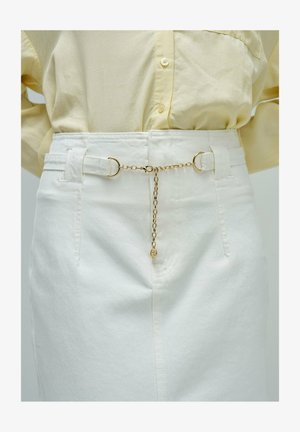 Primer plano de pantalones blancos de tiro alto con trabillas unidas por una delicada cadena dorada y una camisa amarilla clara abotonada metida por dentro.