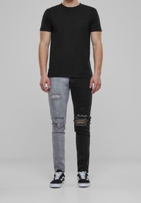 Camisa negra combinada con jeans desgastados que presentan un diseño de color dividido: gris a la izquierda y negro a la derecha, acentuado con costuras visibles.