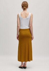 Geel gestructureerde lange rok met een aangesloten tailleband, gecombineerd met een mouwloze witte top. Glanzende zwarte platte sandalen maken de outfit af.