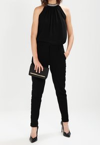 Svart halterneck toppen med samlad tyg och smyckad krage, parat med skräddarsydda svarta skinny jeans och svarta högklackade skor, hållande en kuvertväska.