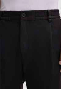 Schwarze Hose aus einem strukturierten Stoff, mit elastischem Bund und Knopfverschluss sowie seitlichen Taschen, Teil eines modernen Designs.