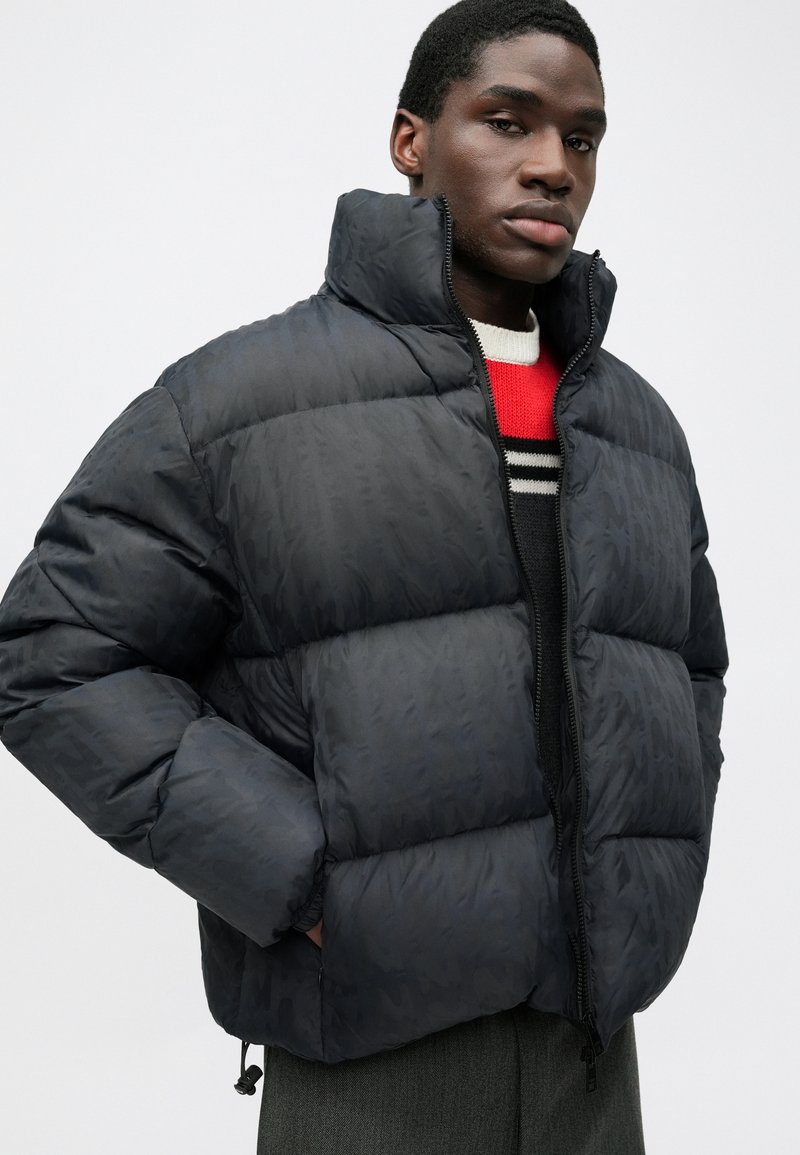 Puffer jacket in dark navy, featuring a high collar, textured fabric, zip closure, and elastic cuffs. Layered over a red and striped sweater.