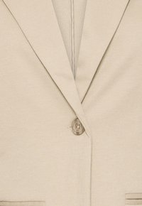 Blazer beige in tessuto strutturato, chiusura con un solo bottone, revers a lancia e due tasche frontali orizzontali visibili nella parte inferiore.