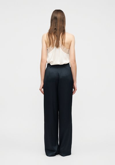 Γυναίκα με μακριά μαλλιά που φοράει λευκό, αμάνικο τοπ με δαντέλα και μαύρο, ψηλόμεσο παντελόνι με φαρδύ πόδι, στέκεται μπροστά σε μονόχρωμο φόντο.