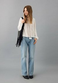 Blusa blanca de manga larga con detalles bordados, combinada con jeans rectos de color azul claro y botines negros, sosteniendo un bolso negro con flecos.