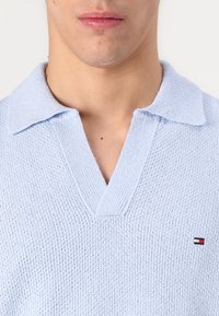 Svetlomodrá pletená polo tričko s V-krkom, textúrovaná látka a malý detail loga na ľavej hrudi.