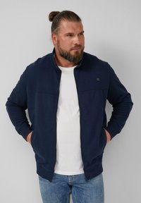 Giacca zip-up blu navy con colletto alto, due tasche frontali e una superficie liscia. Indossata sopra una camicia bianca, abbinata a jeans azzurri chiari.
