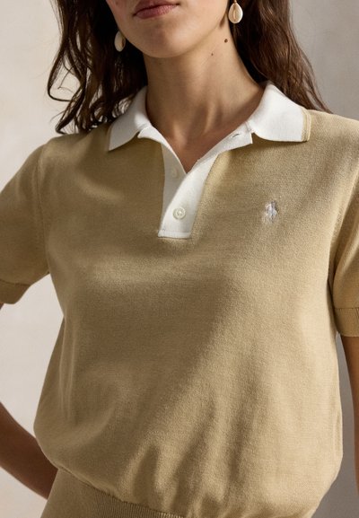 Γυναίκα που φοράει μπεζ κοντομάνικο πλεκτό πόλο πουκάμισο με λευκή γιακά και κουμπότρυπες, σκουλαρίκια με κέλυφος, στέκεται με το ένα χέρι στη μέση.