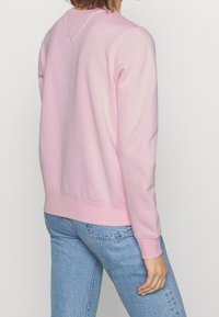 Vaaleanpunainen collegepaita pyöreällä pääntiellä, resoroiduilla hihansuuilla ja helmalla, pehmeällä tekstuurilla ja kolmionmuotoisella selkäyksityiskohdalla, yhdistettynä sinisiin farkkuihin.