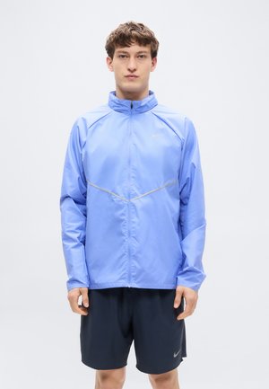 Nuori mies katsomassa suoraan eteenpäin, päällään vaaleansininen vetoketjullinen takki ja mustat urheilushortsit valkoista taustaa vasten.