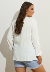 Weißes Langarmoberteil mit strukturierter Oberfläche und lockerer Passform, kombiniert mit hellblauen Shorts. Der Hintergrund ist in einer neutralen, einfarbigen Farbe gehalten.
