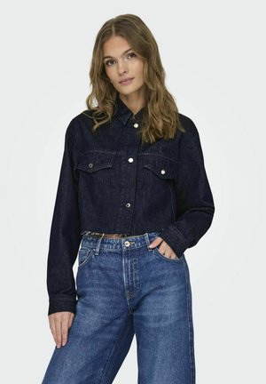 Giacca in denim scuro tagliata con due tasche sul petto, accessori in argento e chiusura a bottone. Abbinata a jeans blu a vita alta.