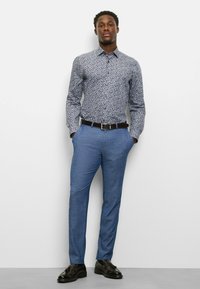 Hemd mit Knopfleiste, verziert mit einem Blumendesign in Blau, Weiß und Rot; kombiniert mit hellblauen Hosen und schwarzen Lederschuhen.