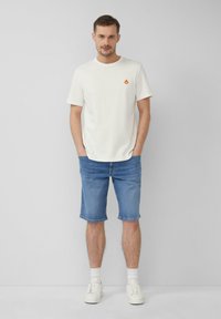 T-shirt en coton blanc avec un petit logo de flammes rouges et jaunes, associé à un short en denim clair. Le modèle porte des baskets blanches et des chaussettes courtes.
