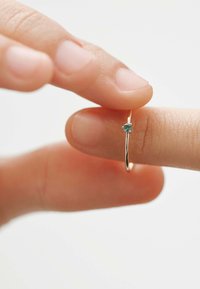 Pendiente de aro dorado con una pequeña gema redonda de tono teal, sostenido entre dos dedos. Superficie metálica lisa, diseño circular sencillo.