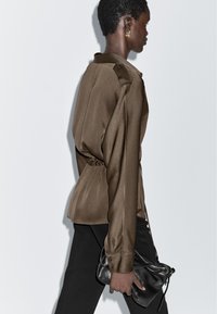 Bruine satijnen blouse met lange mouwen, een geplooide taille en manchetten met knopen, gecombineerd met een zwarte broek en een kleine zwarte handtas.