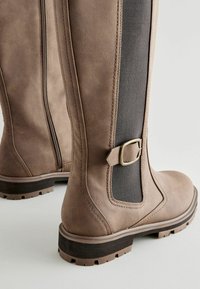Brown knee-high boots made of textured leather with a side zipper and herringbone elastic panel, featuring a metal buckle accent.