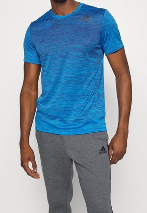 Mann trägt ein blaues kurzärmeliges Sportshirt und graue Adidas-Sweatpants vor weißem Hintergrund.
