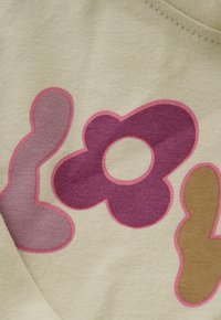 Nahaufnahme von beigem Stoff mit bunten, verspielten Formen: einem mauvefarbenen Buchstaben "L", einer lila Blume mit hohlem Zentrum und einem senfgelben Buchstaben "Y".