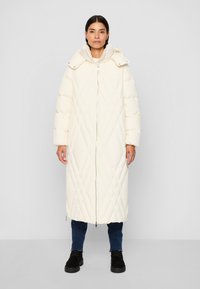 Woman standing wearing a long, quilted cream coat with a high collar, zipped front, dark jeans, and black lace-up boots on a plain background.