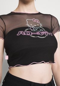 Svart nätcroppad topp med rosa vågiga kanter, med en Hello Kitty-grafik och japansk text i klar brodyr.
