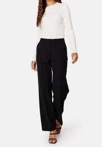 Weiße langärmlige Bluse aus strukturiertem Stoff, kombiniert mit hochgeschnittenen schwarzen weit geschnittenen Hosen. Schwarze Riemenabsätze zeigen die Zehen.