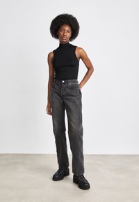 Mujer con peinado afro, vistiendo un cuello de tortuga sin mangas negro, jeans oscuros de talle alto y zapatos de plataforma negros, de pie contra una pared lisa.