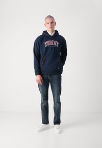 Námornícka mikina s bielym a červeným textom "TOMMY JEANS", predným vreckom a sťahovacou kapucňou. Kombinovaná s tmavými džínami a bielymi teniskami.