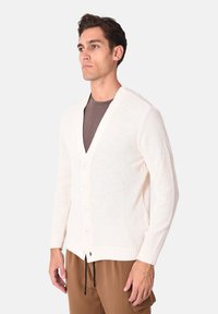 Cardigan bianco in maglia con scollo a V, maniche lunghe e sei bottoni frontali. Tessuto strutturato e vestibilità rilassata, abbinato a pantaloni marroni.