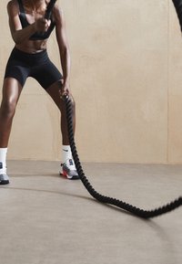 Tenue de sport noire portée par une personne agrippant une corde de combat épaisse et noire. En arrière-plan, un mur texturé de couleur brun clair et un sol gris.