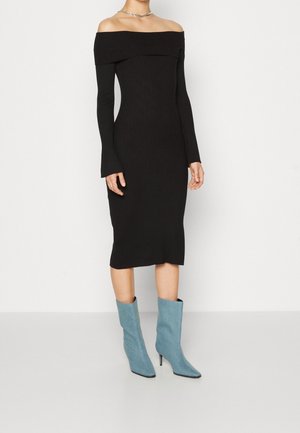 Femme portant une robe midi côtelée noire à épaules dénudées et des bottes à talons pointues bleu clair, se tenant devant un fond blanc uni.