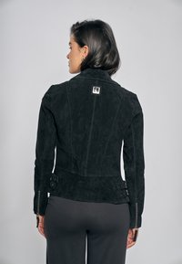 Černá semišová bunda s přiléhavým střihem, límcem a nastavitelnými bočními pásky. Výrazné zipy na manžetách a značková nášivka na zádech.