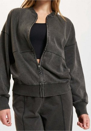 Kvinde iført mørkegrå lynlåsjakke over sort crop top og matchende mørkegrå bukser, stående med armene afslappet ned langs siden.