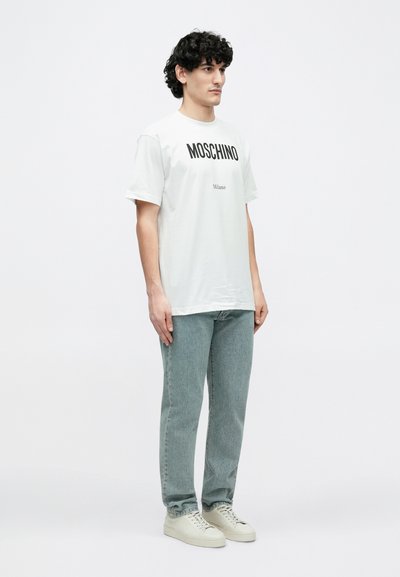 Biały bawełniany t-shirt z nadrukiem "MOSCHINO" i "Milano" w pogrubionych czarnych literach. Noszony z jasnoniebieskimi dżinsami i beżowymi trampkami.