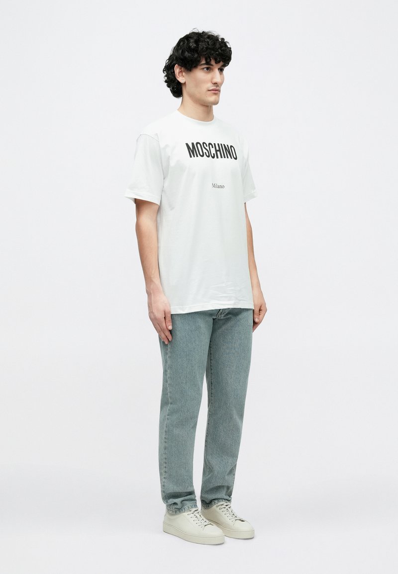 Hvid bomuld t-shirt med "MOSCHINO" og "Milano" trykt i fede sorte bogstaver. Båret med lyseblå denimjeans og beige sneakers.