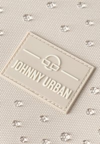 Svetlo béžová látková taška s textúrovanými bodkami, s výraznou bielou embosovanou nálepkou so nápisom "JOHNNY URBAN" v obdĺžnikovom tvare.