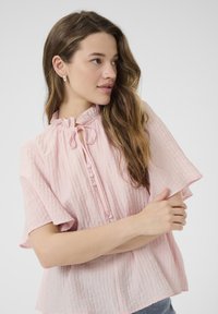 Light pink blouse made of textured fabric, featuring short flutter sleeves, a high neck with a tie, and button detail down the front.