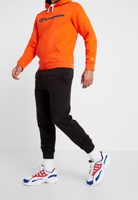 Sudadera naranja con el logo negro de "Champion", pantalones de chándal negros y zapatillas blancas con acentos rojos y azules y suelas gruesas.