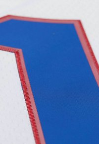 Weißes Sporttrikot mit einer großen blauen Nummer "1", die rot umrandet ist. Der Stoff weist ein strukturiertes Muster mit Perforationen für Atmungsaktivität auf.