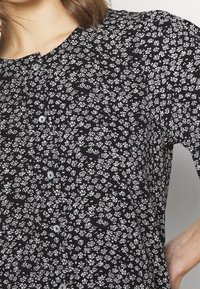 Blouse noire à petit motif floral blanc, fermetures à boutons et encolure ronde. Tissu doux et léger avec une finition texturée.