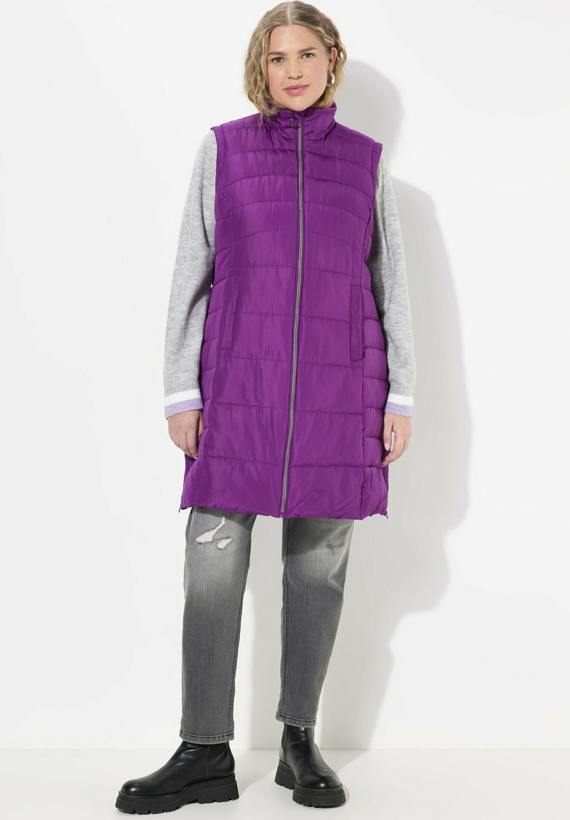 Lila gepolsterte Weste mit hohem Kragen, vorderem Reißverschluss und zwei seitlichen Taschen, getragen über einem grauen Pullover und kombiniert mit verwaschenen grauen Jeans und schwarzen Stiefeln.