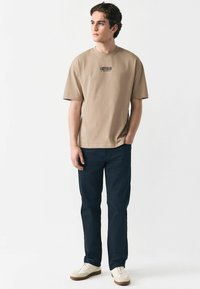 Camiseta beige de manga corta con la palabra "AMSTERDAM" impresa en negro, combinada con pantalones oscuros y zapatillas claras, sobre un fondo liso.