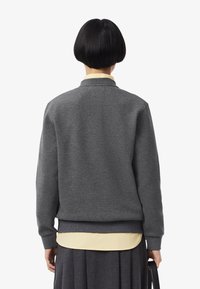 Persona con un taglio di capelli bob corto e nero, che indossa un maglione grigio su una camicia beige e una gonna grigio scuro, tenendo una borsa nera al suo fianco.