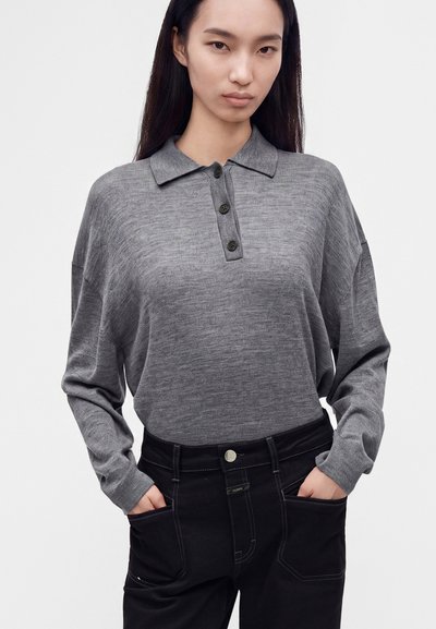 Jeune femme aux longs cheveux foncés porte un polo gris à manches longues et un pantalon noir, les mains dans les poches, sur un fond uni.