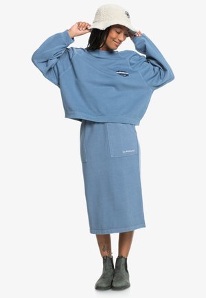 Completo composto da felpa blu corta e gonna lunga in morbido cotone, caratterizzato da un dettaglio logo frontale. Abbinato a un cappello a secchiello crema e stivali scuri.