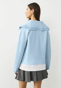 Jasnoniebieski krótki sweter z szerokim kołnierzem marynarskim zdobionym niebieskimi paskami, zestawiony z białą koszulą i szarą plisowaną spódnicą.