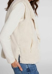 Beige väst i fuskpäls med mjuk textur, öppen framdel och sidofickor; lager på en krämfärgad långärmad skjorta, kombinerad med blå jeans.