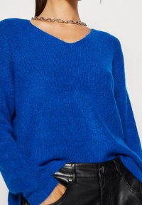 Blå stickad tröja med V-ringning, avslappnad passform och ribbad struktur. Matchas med svarta skinnbyxor och ett silverkedjehalsband.