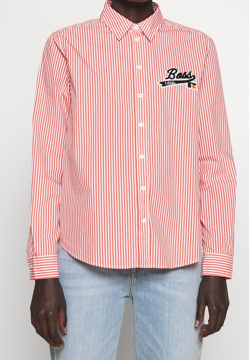 Chemise rayée verticale rouge et blanche à boutons avec écusson « Boss Athletic » sur la poitrine gauche, portée avec un jean bleu clair.